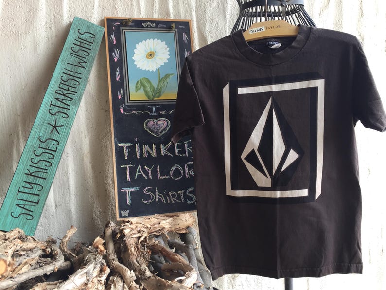 Pu&ograve; includere: Una maglietta marrone con un logo a forma di diamante bianco. Uno sfondo con una lavagna nera che dice "Tinker Taylor T Shirts".
