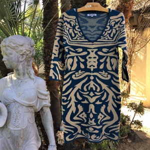 May include: A teal blue and beige patterned sweater dress with a V-neck and three-quarter sleeves. The dress is hanging on a wooden hanger with a "Taylor" tag.