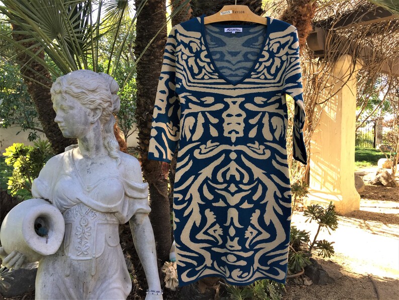 May include: A teal blue and beige patterned sweater dress with a V-neck and three-quarter sleeves. The dress is hanging on a wooden hanger with a "Taylor" tag.