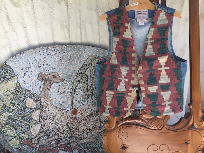 K&ouml;nnte beinhalten: Eine Jeansweste mit einem farbenfrohen geometrischen Muster auf der Vorderseite. Die Weste hat einen braunen Lederriemen und einen Metallknopfverschluss. Die Weste ist von "Jonah & Smith" hergestellt und hat ein "Vintage Style" Etikett.