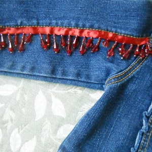 May include: Close-up of a denim jacket with red beaded fringe and a red ribbon trim. The jacket features an American flag patch on the sleeve. The denim is a medium blue wash.