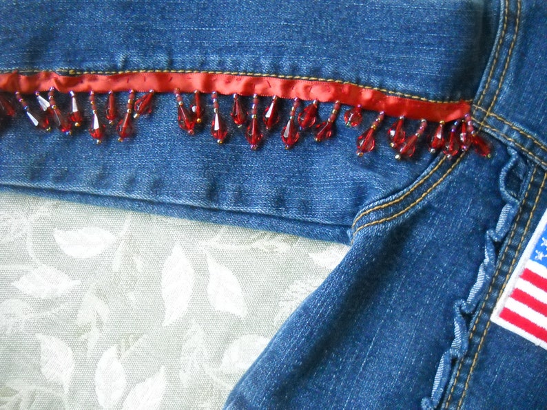 May include: Close-up of a denim jacket with red beaded fringe and a red ribbon trim. The jacket features an American flag patch on the sleeve. The denim is a medium blue wash.