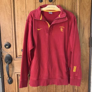 May include: A red long-sleeve sweatshirt with a yellow collar and accents. The Nike logo is visible on the chest, and the letters "USC" are printed on the sleeve. The sweatshirt is hanging on a wooden door.