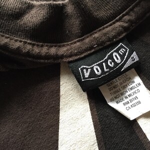 Pu&ograve; includere: Una maglietta a righe marroni e bianche con un'etichetta Volcom nera. L'etichetta presenta il testo "Volcom" in lettere bianche e include le istruzioni per la cura.