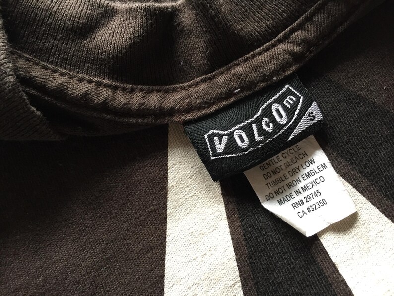 Pu&ograve; includere: Una maglietta a righe marroni e bianche con un'etichetta Volcom nera. L'etichetta presenta il testo "Volcom" in lettere bianche e include le istruzioni per la cura.