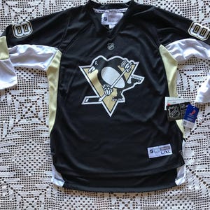 May include: Black and gold hockey jersey with the Pittsburgh Penguins logo on the front. The jersey has white and gold accents on the sleeves and shoulders. The number "00" is on the sleeves. The jersey is a size L/XL.