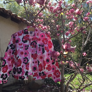 May include: A white denim jacket with a pink and black floral print. The jacket is hanging on a hanger in front of a tree with pink flowers.