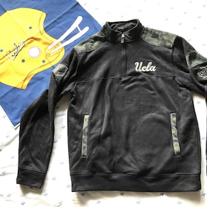May include: Black and green camouflage quarter-zip sweatshirt with a white "UCLA" logo on the front.