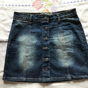 May include: A dark blue denim skirt featuring a high-waisted design with a vertical row of six metallic buttons down the center front. The fabric displays a faded, light-wash effect on the front panels and includes visible stitching details.