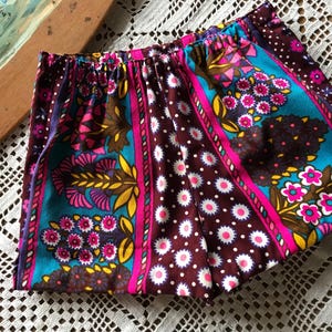 May include: Colorful vintage shorts with an elastic waistband. The shorts feature a vibrant floral pattern in teal, pink, yellow, and brown. The fabric has a retro aesthetic, perfect for a summer outfit.
