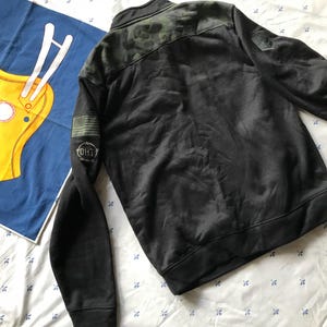Puede incluir: Chaqueta negra y verde con estampado de camuflaje, cremallera y un parche negro con texto blanco que dice "OH1T" en la manga izquierda.