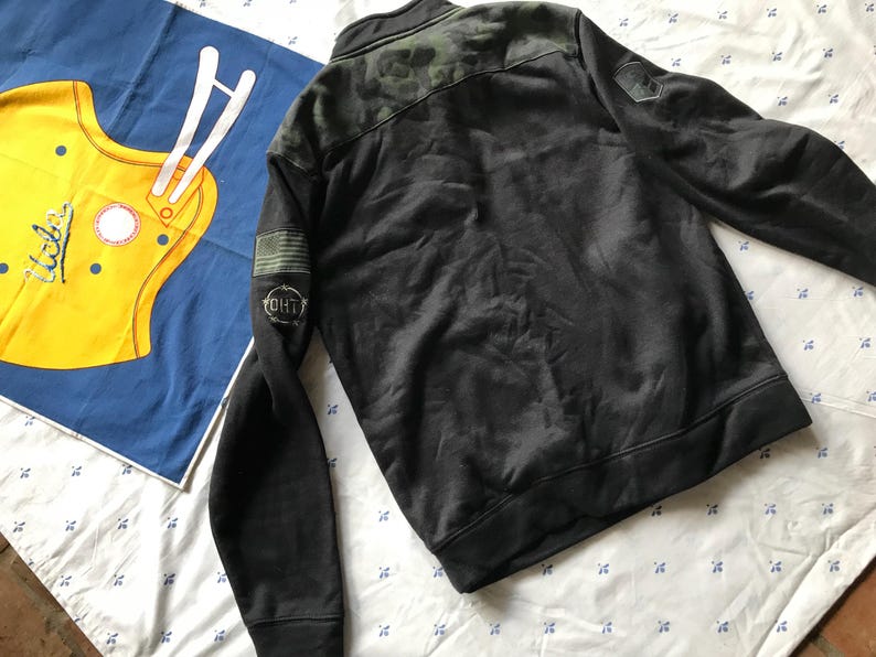 Puede incluir: Chaqueta negra y verde con estampado de camuflaje, cremallera y un parche negro con texto blanco que dice "OH1T" en la manga izquierda.