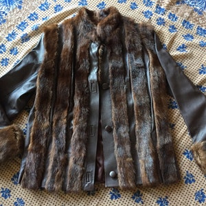 May include: A brown fur and leather jacket with a button-up front. The jacket has fur panels down the front and on the cuffs, with leather sleeves and a collar. The jacket is displayed on a patterned fabric.