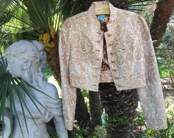 May include: A cropped gold and white paisley patterned jacket with gold buttons and a leopard print lining. The jacket is hanging on a wooden hanger and is displayed against a backdrop of green foliage.