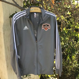 May include: Gray Adidas track jacket with white stripes on the sleeves and the Adidas logo on the chest. The jacket has a full zipper and a Dynamo logo on the chest.