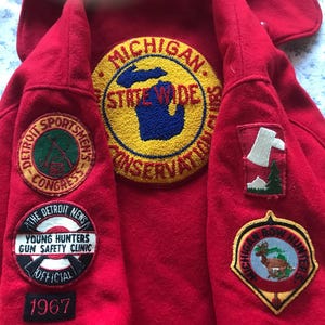 May include: A red jacket with multiple embroidered patches. Patches include "Michigan State Wide Conservation", "Detroit Sportsmen's Congress", "Young Hunters Gun Safety Clinic Official 1967", and "Michigan Bow Hunters".