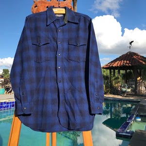 May include: A blue and black plaid button-down shirt with two chest pockets. The long-sleeved shirt is hanging on a wooden hanger outdoors. The shirt is made of a soft, comfortable material.