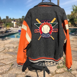 May include: A gray baseball jacket with orange leather sleeves and white stripes. The back features a "Louisville Slugger" patch with a baseball and crossed bats. The jacket has a striped collar, cuffs, and hem.