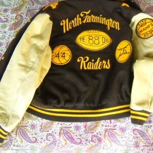 May include: A black and yellow varsity jacket with leather sleeves. The jacket has the text "North Farmington Raiders" and "City Champs 93-94" embroidered on it. There are also patches with the numbers "44", "88", and "76" on the jacket.