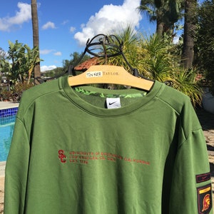May include: Green sweatshirt with a red embroidered logo that says "University of Southern California Los Angeles, CA 90089 California" and a Nike swoosh on the chest. The sweatshirt has a red and gold patch on the sleeve with a Trojan warrior logo.