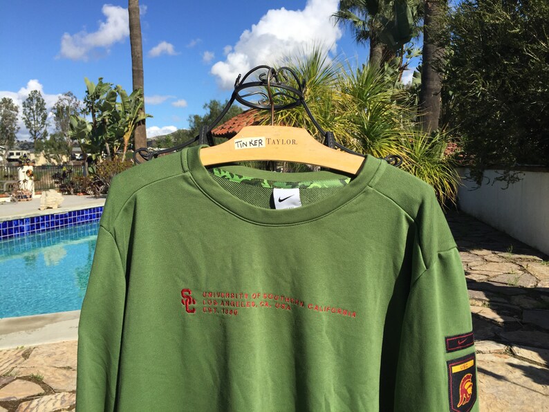 May include: Green sweatshirt with a red embroidered logo that says "University of Southern California Los Angeles, CA 90089 California" and a Nike swoosh on the chest. The sweatshirt has a red and gold patch on the sleeve with a Trojan warrior logo.