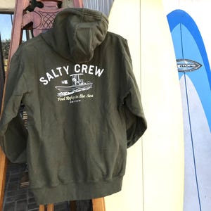 May include: Olive green hooded sweatshirt with the text "SALTY CREW" and a boat graphic on the back. The sweatshirt also has the text "Find Refuge in the Sea" and "SINCE 2013". The sweatshirt is hanging on a wooden ladder next to a surfboard.