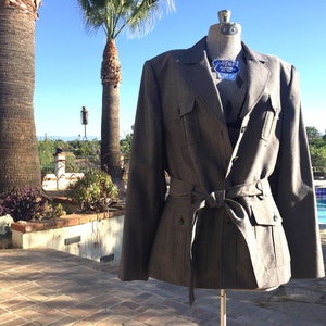 May include: A gray, belted jacket with a double-breasted closure and flap pockets. The jacket is on a mannequin in front of a swimming pool.