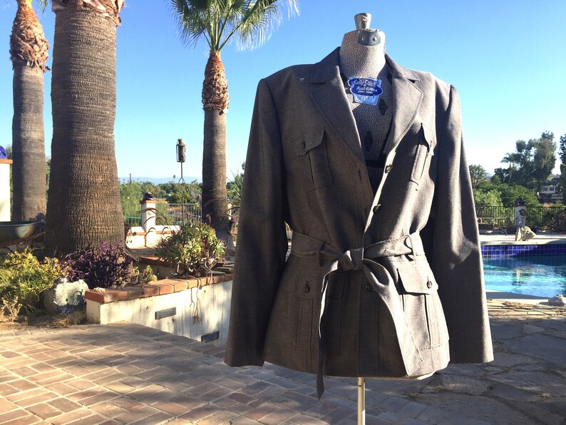 May include: A gray, belted jacket with a double-breasted closure and flap pockets. The jacket is on a mannequin in front of a swimming pool.