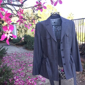 May include: A gray blazer with a belt and two flap pockets. The blazer is on a mannequin and has a white label with text that is not visible.