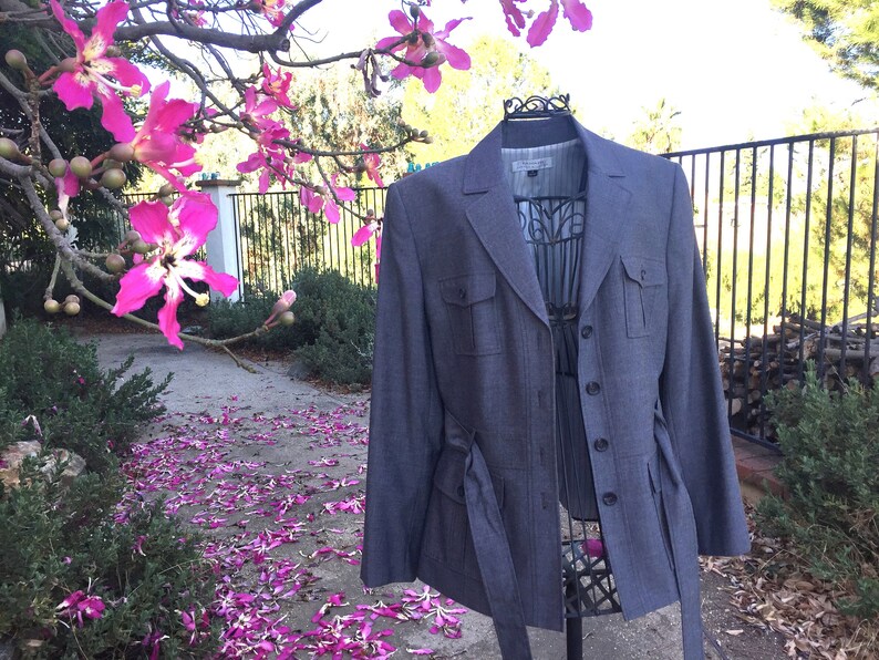 May include: A gray blazer with a belt and two flap pockets. The blazer is on a mannequin and has a white label with text that is not visible.