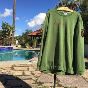 May include: Green long-sleeved sweatshirt with a small embroidered logo on the left chest and a larger patch on the right sleeve. The sweatshirt has a crew neck and ribbed cuffs.