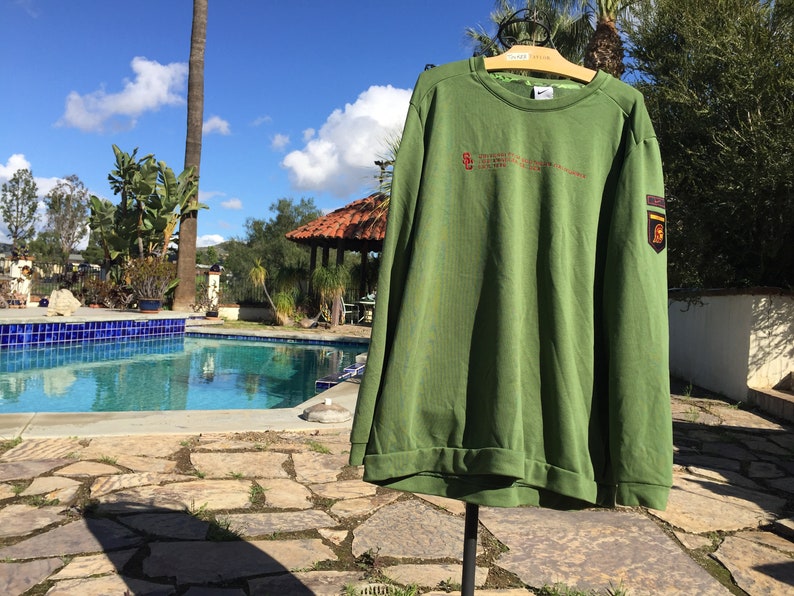 May include: Green long-sleeved sweatshirt with a small embroidered logo on the left chest and a larger patch on the right sleeve. The sweatshirt has a crew neck and ribbed cuffs.