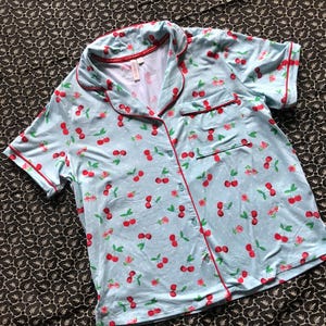 May include: Light blue pajama top with a cherry print. The short-sleeved shirt has a button-down front, a chest pocket, and red trim. The cherry pattern includes red cherries and green leaves on a light blue background.