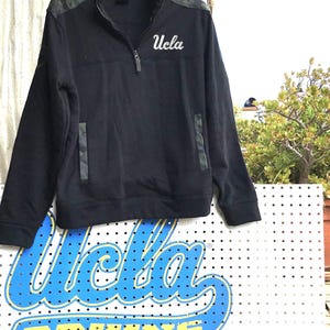 Puede incluir: Sudadera negra de cremallera de un cuarto con estampado de camuflaje en los hombros y un "UCLA" blanco bordado en el pecho.