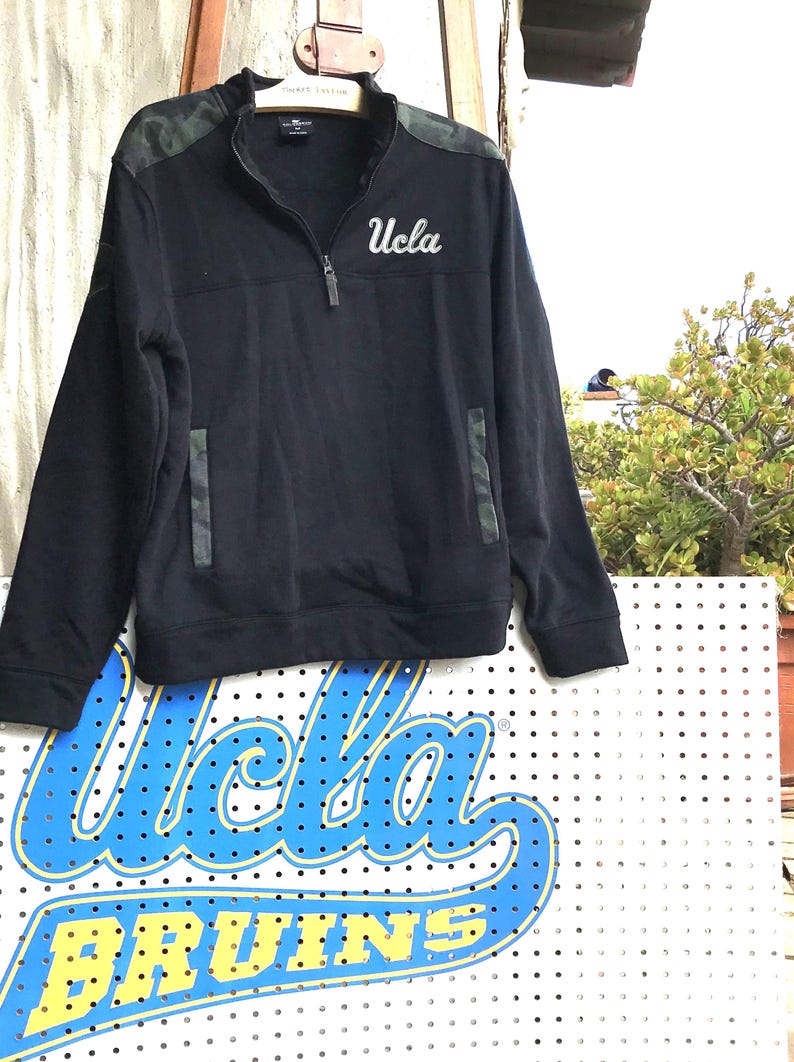 Puede incluir: Sudadera negra de cremallera de un cuarto con estampado de camuflaje en los hombros y un "UCLA" blanco bordado en el pecho.