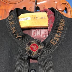 May include: Close-up of a black jacket with gold embroidered text reading "D.K.N.Y." and "Est. 1989" on the collar. The jacket features a decorative pin with red and gold accents. A wooden hanger is visible.