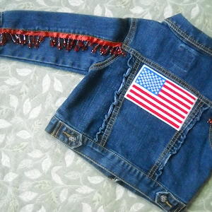 May include: A blue denim jacket with red beaded fringe along the sleeves and a patch of the American flag on the back. The jacket has a classic collar, button closures, and decorative stitching.