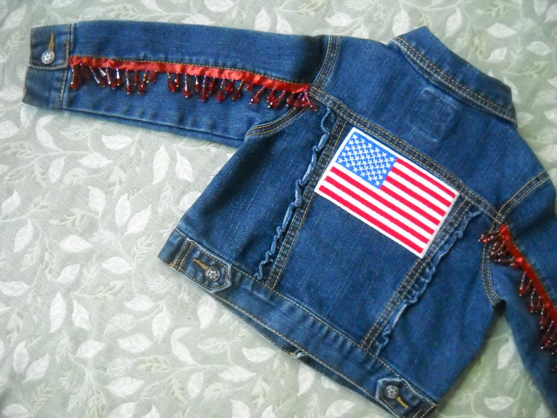 May include: A blue denim jacket with red beaded fringe along the sleeves and a patch of the American flag on the back. The jacket has a classic collar, button closures, and decorative stitching.