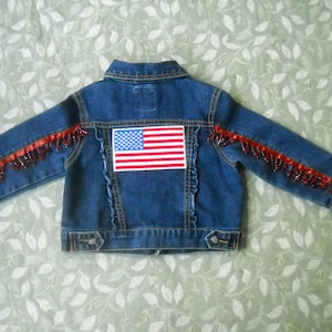 May include: A blue denim jacket with an American flag patch on the back. The sleeves are adorned with red beaded trim. The jacket has a classic collar, button closures, and a decorative ruffled seam.