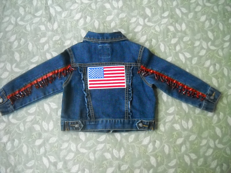 May include: A blue denim jacket with an American flag patch on the back. The sleeves are adorned with red beaded trim. The jacket has a classic collar, button closures, and a decorative ruffled seam.