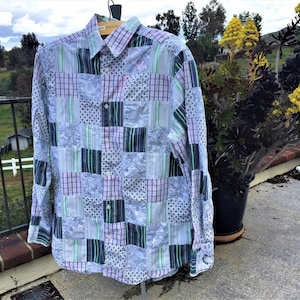 May include: A patchwork button-down shirt with a variety of patterns, including plaid, stripes, and floral designs. The shirt features a color palette of blue, lavender, green, and white. The long-sleeved shirt is hanging on a wooden hanger.