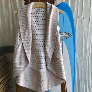 May include: A light beige open-front vest with a textured knit design. The vest features a shawl collar and a geometric pattern on the front panel. It is displayed on a wooden hanger.