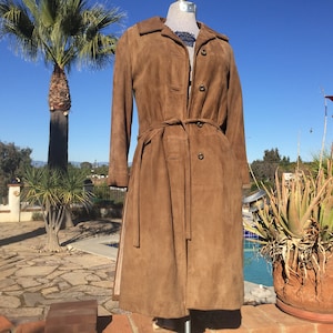 May include: A long brown suede coat with a belt and button closure. The coat is on a mannequin in front of a background of a pool and palm trees.