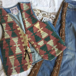 K&ouml;nnte beinhalten: Eine Jeansweste mit einem farbenfrohen geometrischen Muster und einem einzigen Knopfverschluss. Die Weste wird mit einer blauen Jeans mit braunen Lederb&auml;ndern an den Seiten kombiniert.