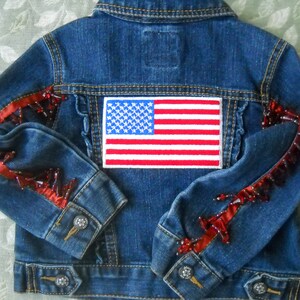 May include: A blue denim jacket with an American flag patch on the back. The sleeves are embellished with red ribbon and red beads. The jacket has silver button closures.