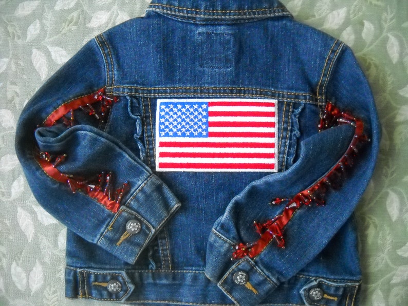 May include: A blue denim jacket with an American flag patch on the back. The sleeves are embellished with red ribbon and red beads. The jacket has silver button closures.