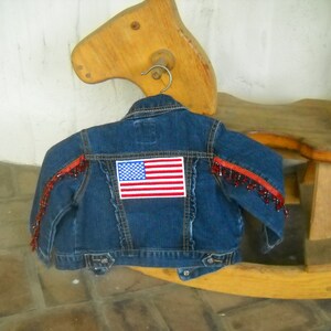 May include: A blue denim jacket with an American flag patch on the back. The jacket has red beaded fringe along the shoulders. The jacket is hanging on a metal hanger.