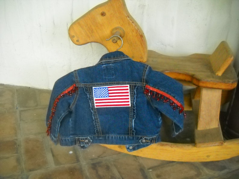 May include: A blue denim jacket with an American flag patch on the back. The jacket has red beaded fringe along the shoulders. The jacket is hanging on a metal hanger.