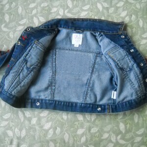 May include: A light blue denim jacket with a classic collar and button closures. The jacket features two front pockets and decorative red accents on the sleeves. The interior lining is a lighter shade of blue.