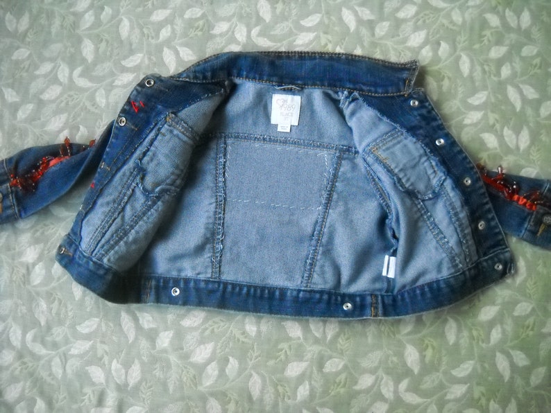 May include: A light blue denim jacket with a classic collar and button closures. The jacket features two front pockets and decorative red accents on the sleeves. The interior lining is a lighter shade of blue.